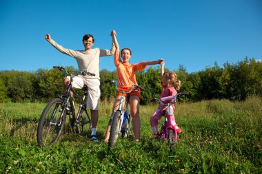 Parents with the daughter on bicycles in park a sunny day. Have clipart