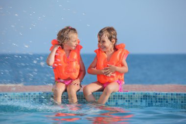 Two little girls in lifejackets sitting on ledge pool on resort, clipart