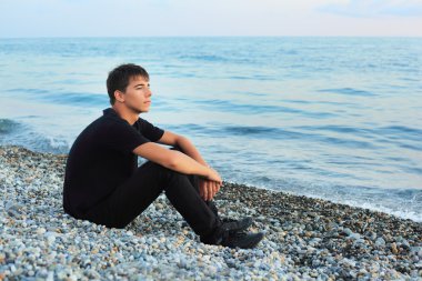 Sitting teenager boy on stone seacoast, Looking afar clipart