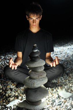 Teenager boy meditating near pyramid from pebble on stony seacoa clipart