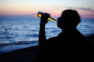 Silhouette teenager boy drinking beer on stone seacoast in eveni clipart