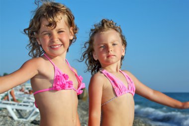 Two pretty little girls on beach near sea, placed hands in sides clipart