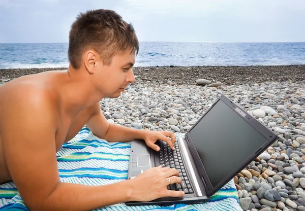 Junger Mann liegt am steinigen Strand und schaut in den Laptop-Bildschirm — Stockfoto