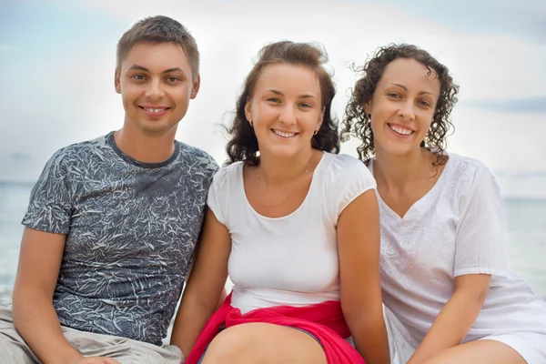 Homem sorridente e duas jovens mulheres bonitas sentadas na praia — Fotografia de Stock
