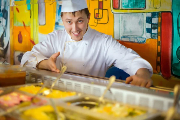 Cocinero alegre en uniforme cerca del mostrador con la comida —  Fotos de Stock