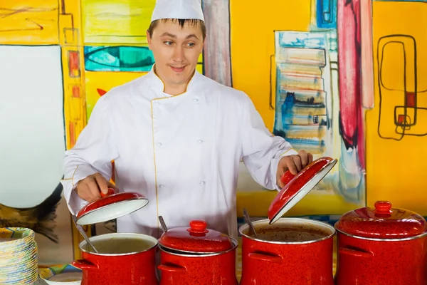 Alegre cocinero en uniforme cerca de sartenes rojas en restaurante público —  Fotos de Stock