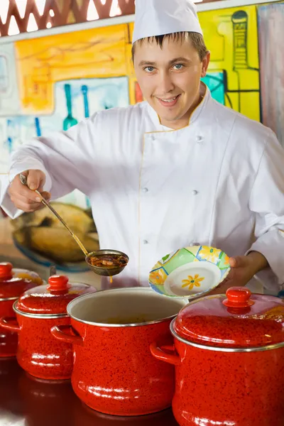 Cheerful cook in uniform spoon imposes soup in plate in public c — Stock Photo, Image