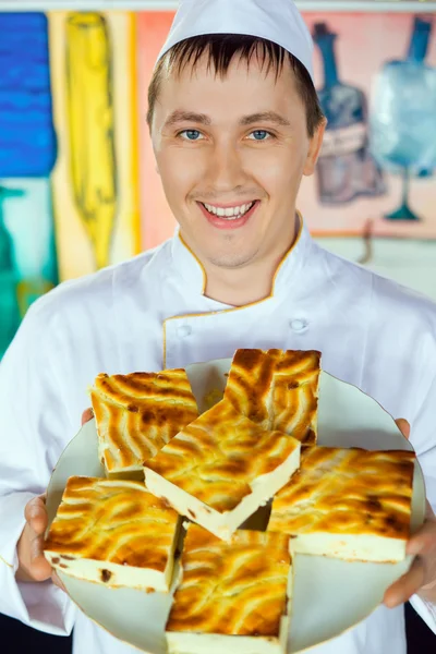 Cheerful cook in uniform holding cheese baked pudding on dish — Stock Photo, Image