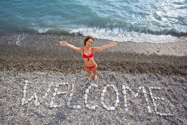 Inschrift aus Steinen willkommen an der steinigen Küste, junge Frau hebt ab — Stockfoto