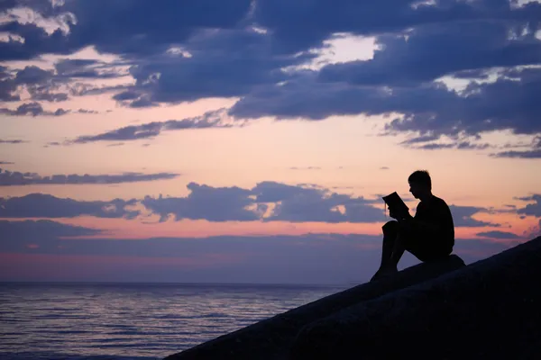 Силует хлопець сидить на розбитій воді ввечері біля моря, читає — стокове фото
