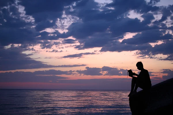 Silhouette srác ül a hullámtörő este a tenger közelében, olvasás — Stock Fotó