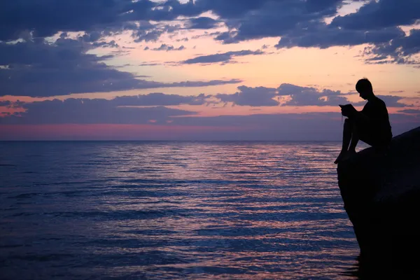 Silueta chlapík sedí na vlnolamu v večer u moře, čtení — Stock fotografie