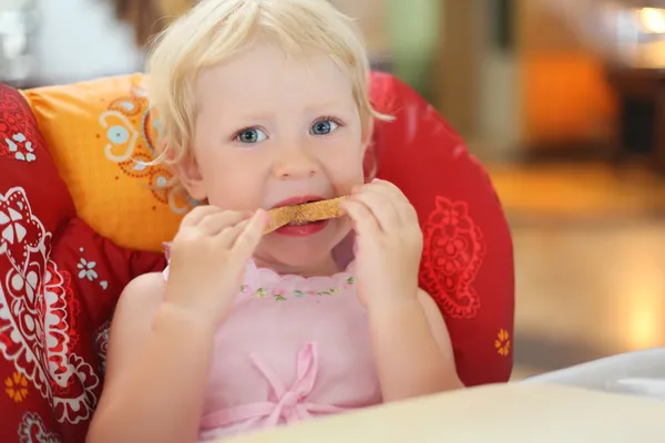 Csinos szőke kislány eszik a kenyeret, egy karosszékben ül — Stock Fotó