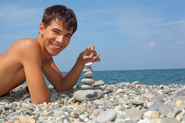 Tiener jongen liggend op steenachtige Zeekust, maakt piramide van pebbl — Stockfoto