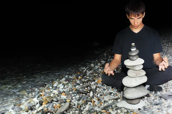 Мальчик-подросток медитирует возле пирамиды из гальки на каменистом морском побережье — стоковое фото