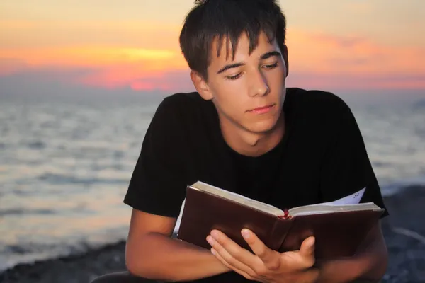 Adolescente menino lê livro sentado na praia à noite — Fotografia de Stock