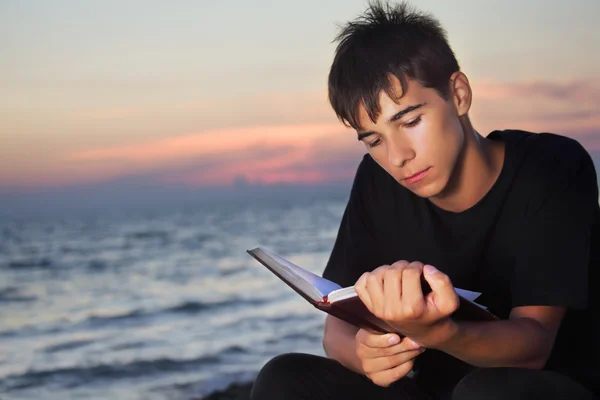Έφηβο αγόρι διαβάζει το βιβλίο κάθεται στην παραλία βράδυ — Φωτογραφία Αρχείου