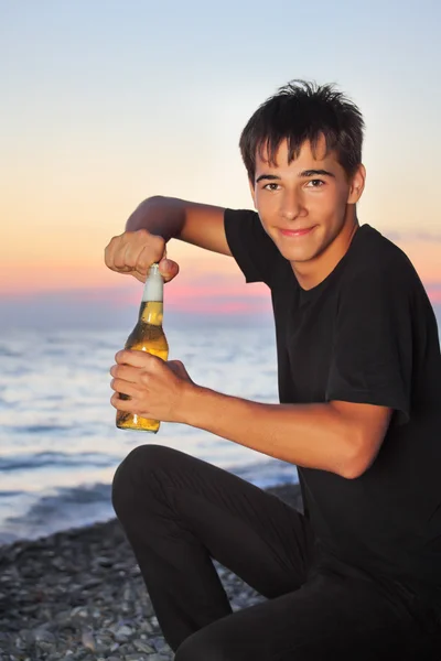 Teenager boy opens beer bottler on stone seacoast in evening — Stockfoto