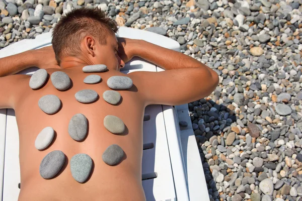 stock image Young man sunbathes on beach with stones on back