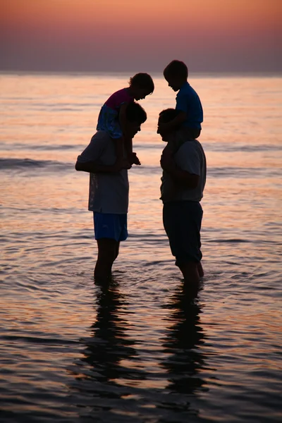 Dois homens no mar com crianças nos ombros ao pôr do sol — Fotografia de Stock