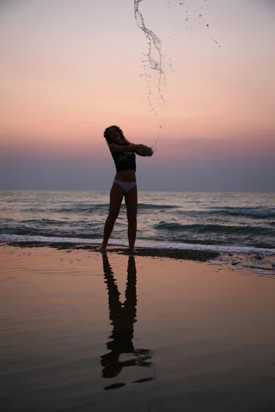 Νεαρή γυναίκα ψεκάζει με νερό στην παραλία στο ηλιοβασίλεμα — Φωτογραφία Αρχείου