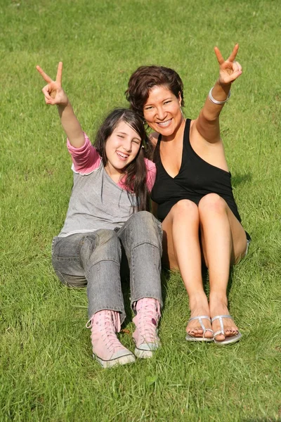 Madre e hija se sientan en la hierba y muestran la señal de la victoria — Foto de Stock