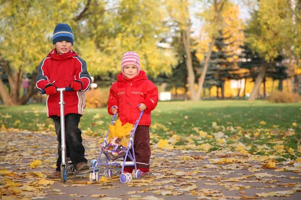 Boy on the scooter and girl with the baby carriage in the park in autumn — Stock Photo, Image