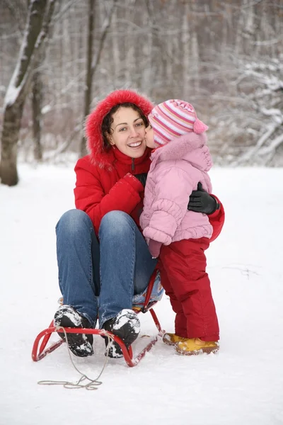 Criança beijo mãe senta-se no trenó no parque no inverno — Fotografia de Stock