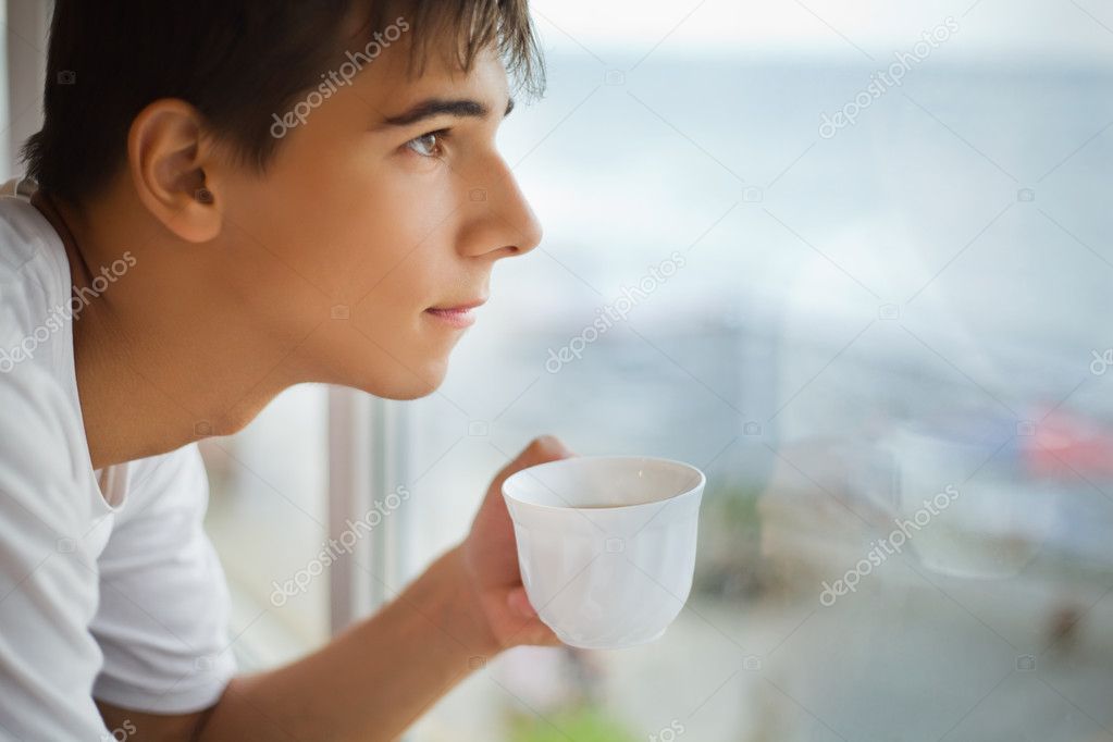 Teenager boy with cup in hand looking out of window in morning,