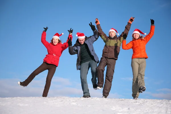 Cuatro amigos saltan en la cima de la colina de nieve en sombreros de Santa Claus —  Fotos de Stock