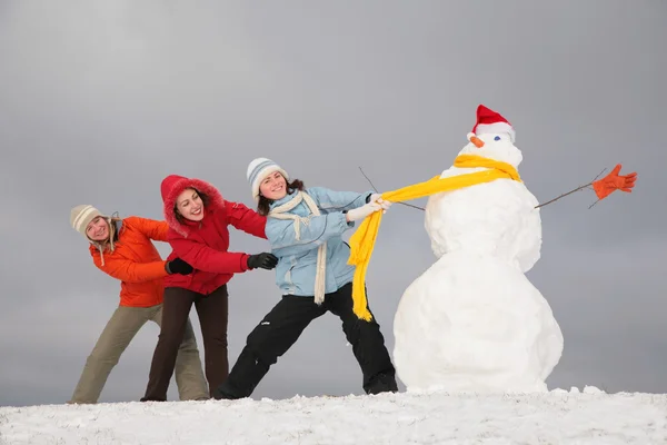 Three young girls pull snowman by yellow scarf — Stock Photo, Image