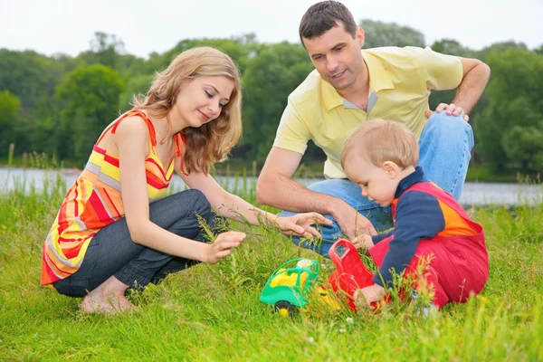Criança senta-se na grama com os pais e brinca com brinquedo — Fotografia de Stock