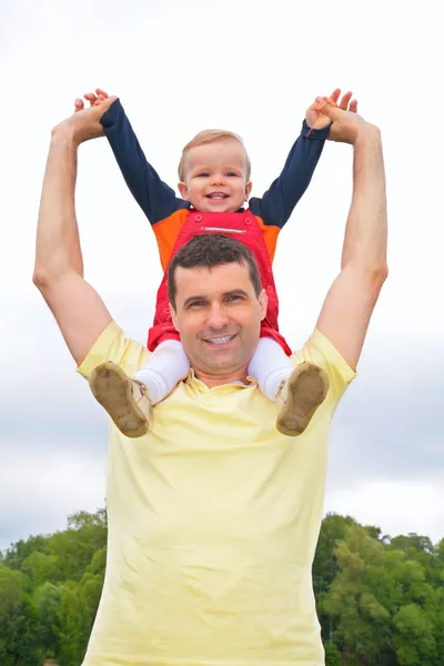 Child sits at father on shoulders with lifted hands outdoors — Stock Photo, Image