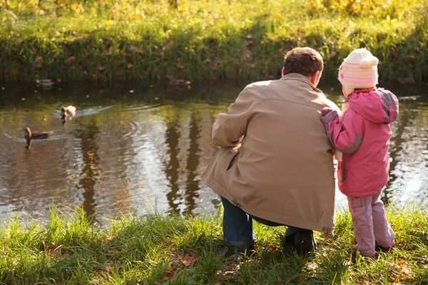 Grandfather with granddaughter in wood in autumn look on ducks in water — Stock Photo, Image