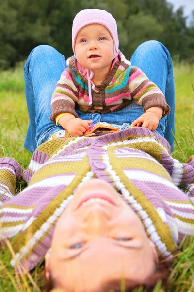 Child sits on mother, who lies on grass — Stock Photo, Image