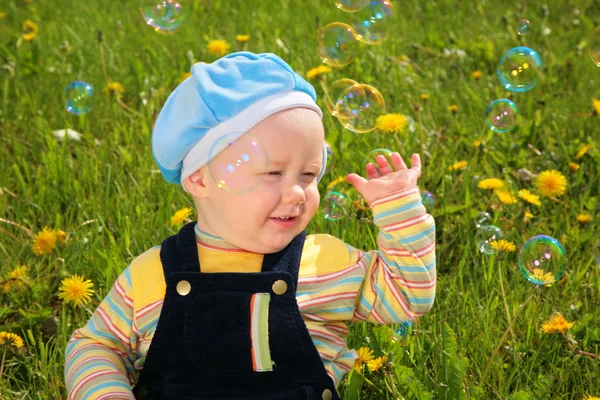 Criança senta-se na grama e olha para bolhas de sabão — Fotografia de Stock