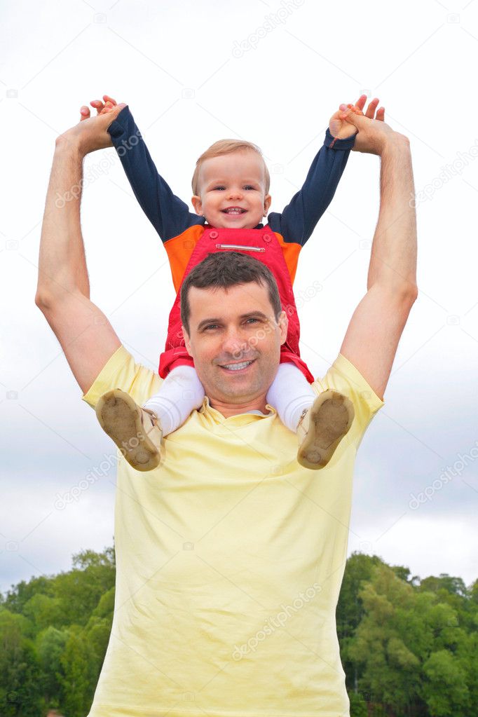Child sits at father on shoulders with lifted hands outdoors