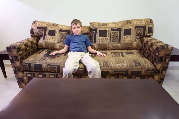Little boy with remote control sitting at brown sofa in room wit — Stock Photo, Image