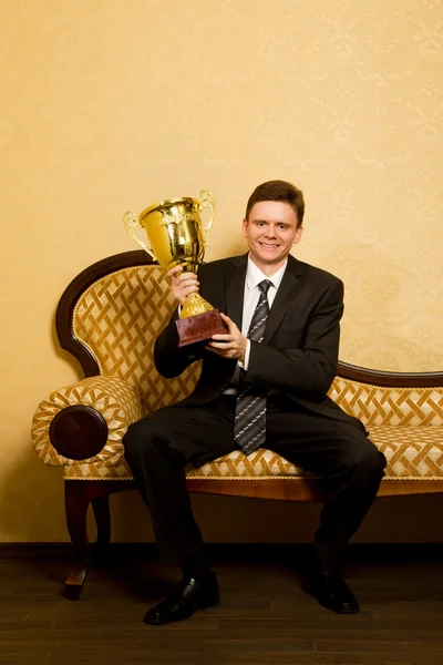 Улыбающийся бизнесмен с кубком победы в руках в костюме сидит на диване — стоковое фото