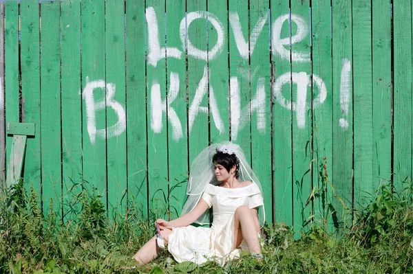 Bruden i brudekjole sitter ved det grønne gjerdet – stockfoto