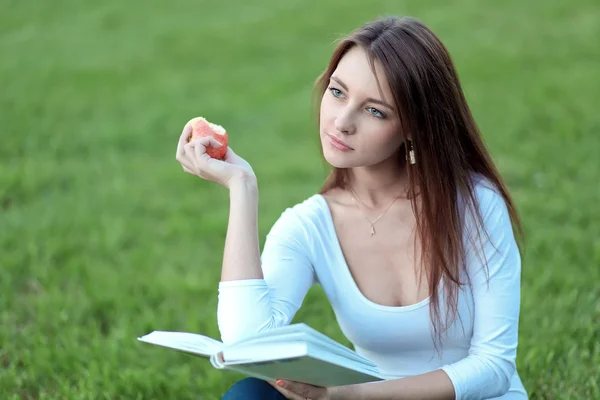 Ładna dziewczyna uczeń na trawniku, czytanie podręcznika i jabłko jedzenie — Zdjęcie stockowe