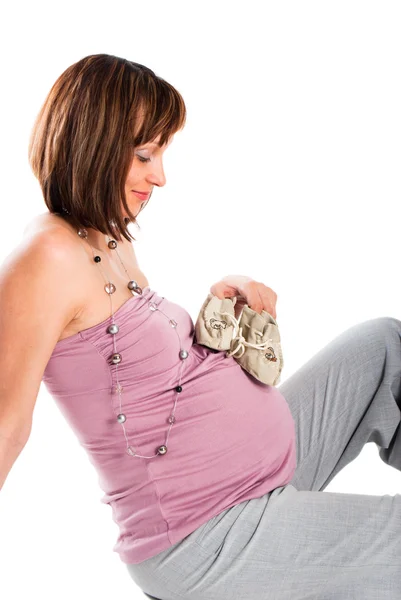 Η έγκυος γυναίκα εξετάζει τις παντόφλες του μωρού, ένα λευκό φόντο — Φωτογραφία Αρχείου