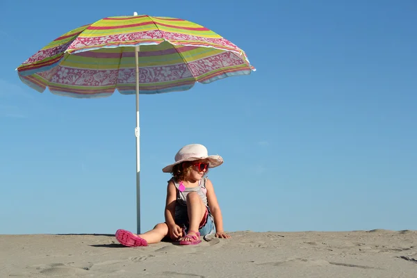 Liten flicka med solglasögon sitter under parasoll — Stockfoto