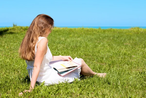 Menina-estudante sentar-se no gramado e lê livro didático — Fotografia de Stock