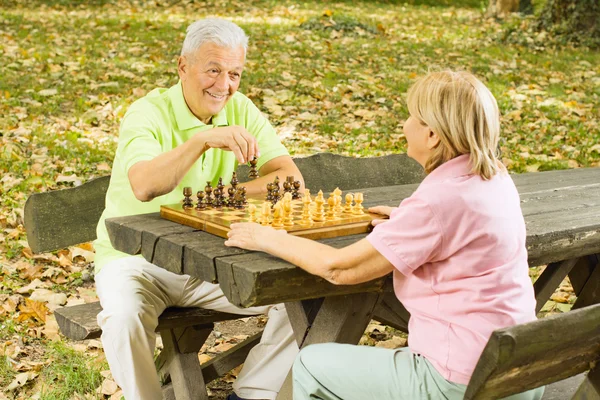 Feliz casal sênior jogar xadrez em um banco de parque — Fotografia de Stock