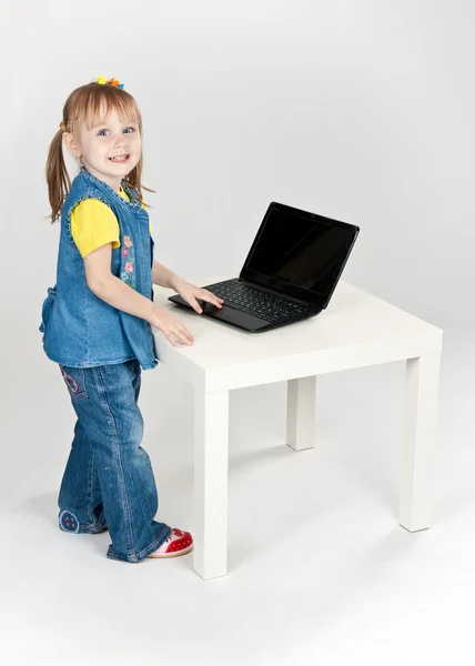 Liten flicka i blå jeans står vid ett bord med en bärbar dator — Stockfoto