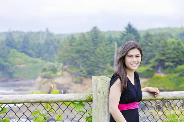Smiling teen girl in black dress standing by ocean shore — Stock Photo, Image