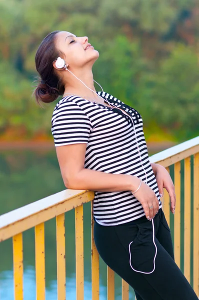 Joven chica atractiva escucha música mientras está de pie en el puente . — Foto de Stock