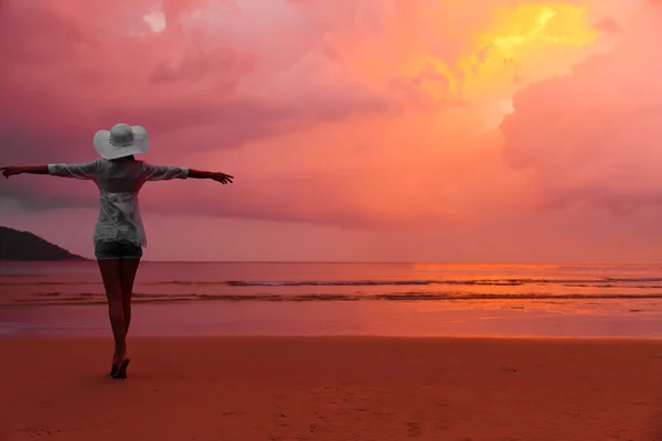 Νεαρή γυναίκα με καπέλο, στέκεται πάνω σε υγρή άμμο και ψάχνει για το κόκκινο sunse — Φωτογραφία Αρχείου