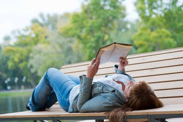 Giovane donna che legge un libro sdraiata sulla panchina — Foto Stock